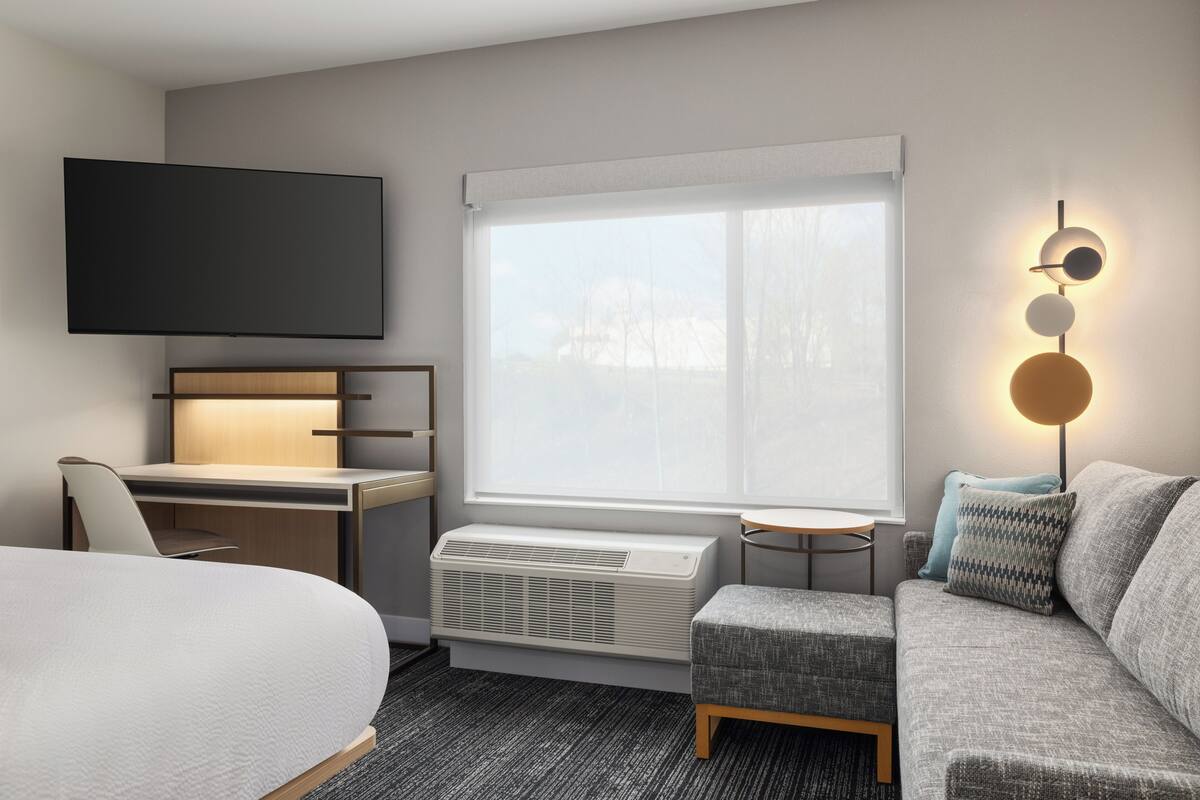 A hotel room with a bed, a desk, a TV, a window with blinds, a heater, a sofa, and a wall-mounted light fixture.