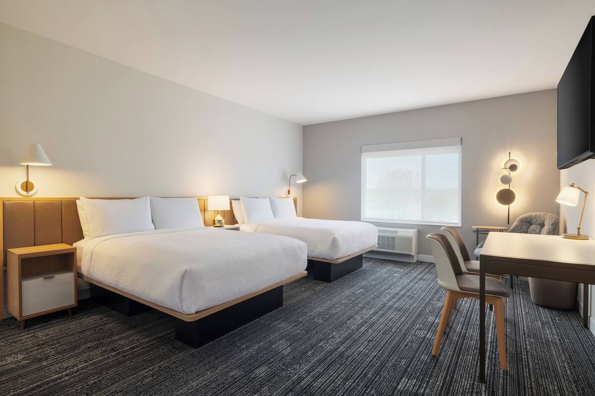 A hotel room with two beds, a desk, a chair, a TV, and a window.