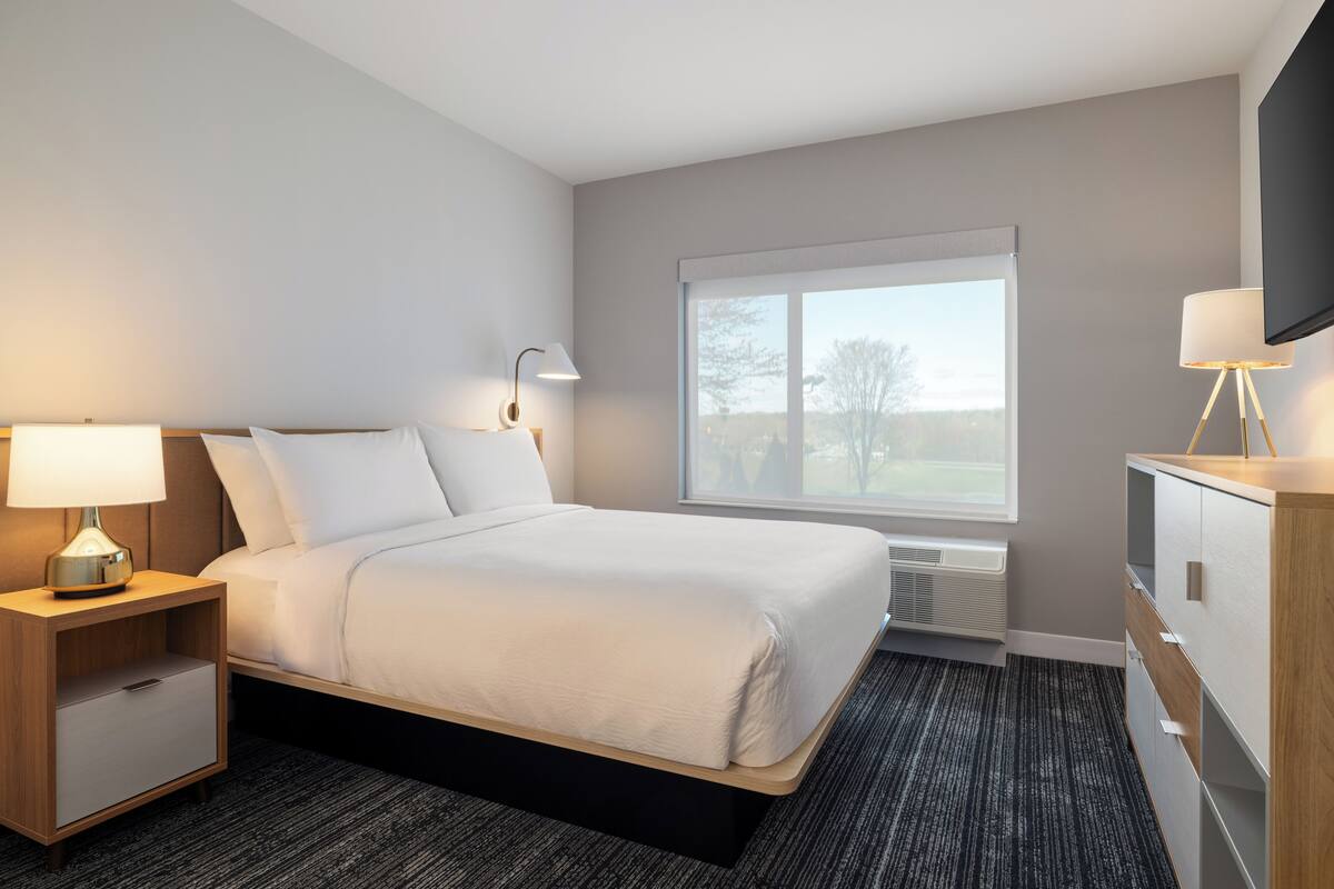 A hotel room with a bed, bedside lamp, TV, and a window with a view.