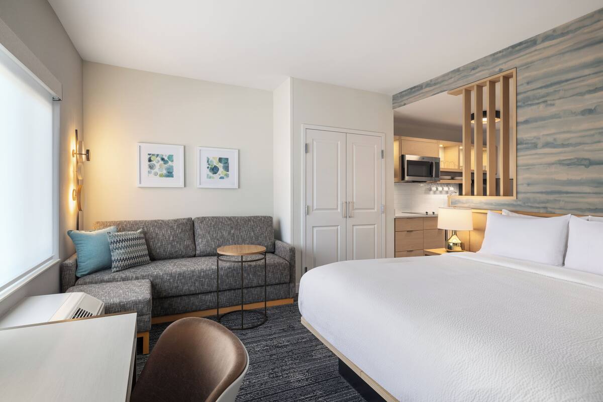 A hotel room with a bed, a sofa, a small table, a chair, a desk, a microwave, a lamp, and artwork on the walls.