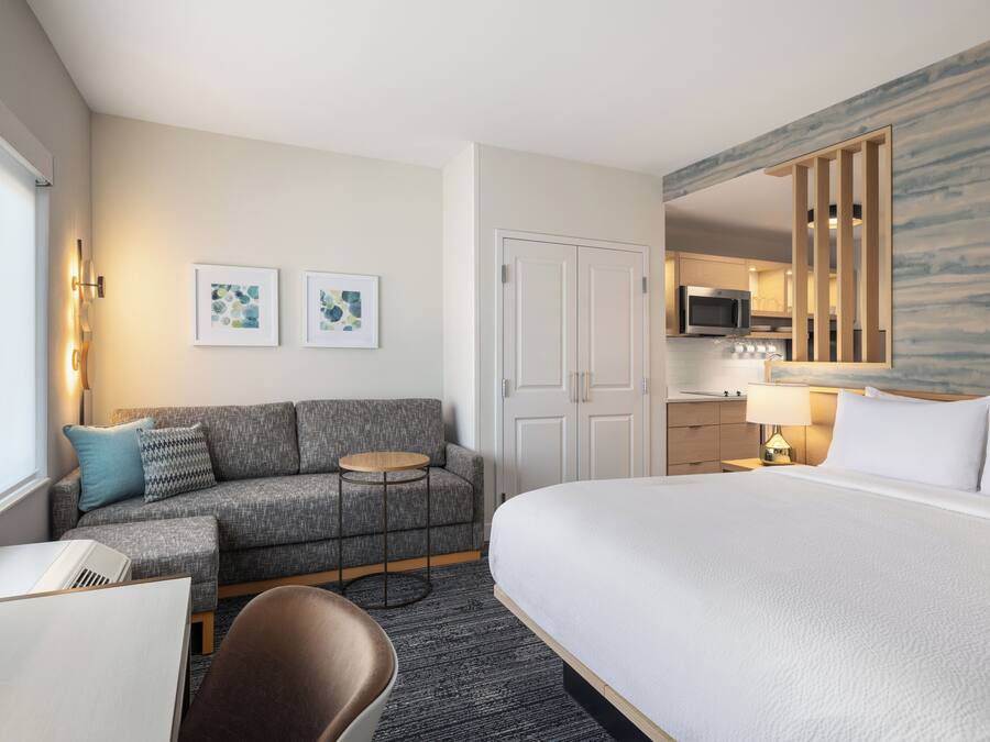Quarto de hotel com cama, sofá, mesa pequena, cadeira, escrivaninha, micro-ondas, luminária e obras de arte nas paredes.