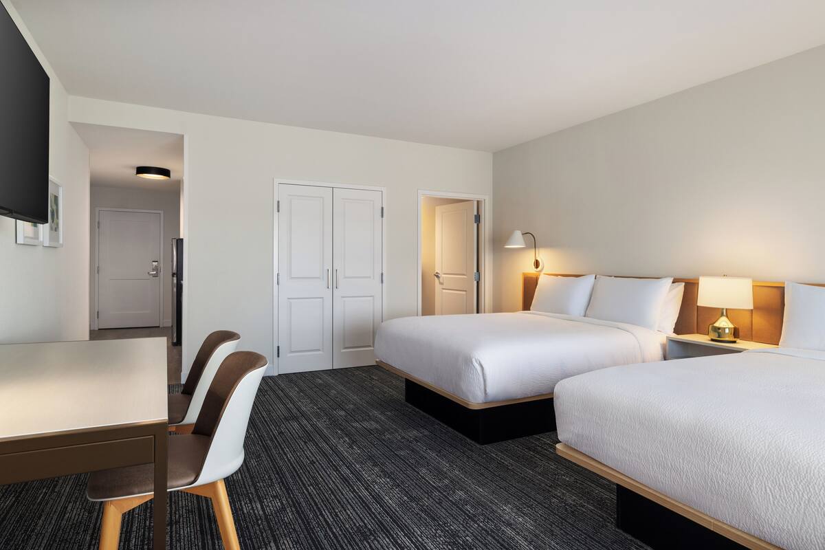 A hotel room with two beds, a desk, chairs, a TV, and a lamp.