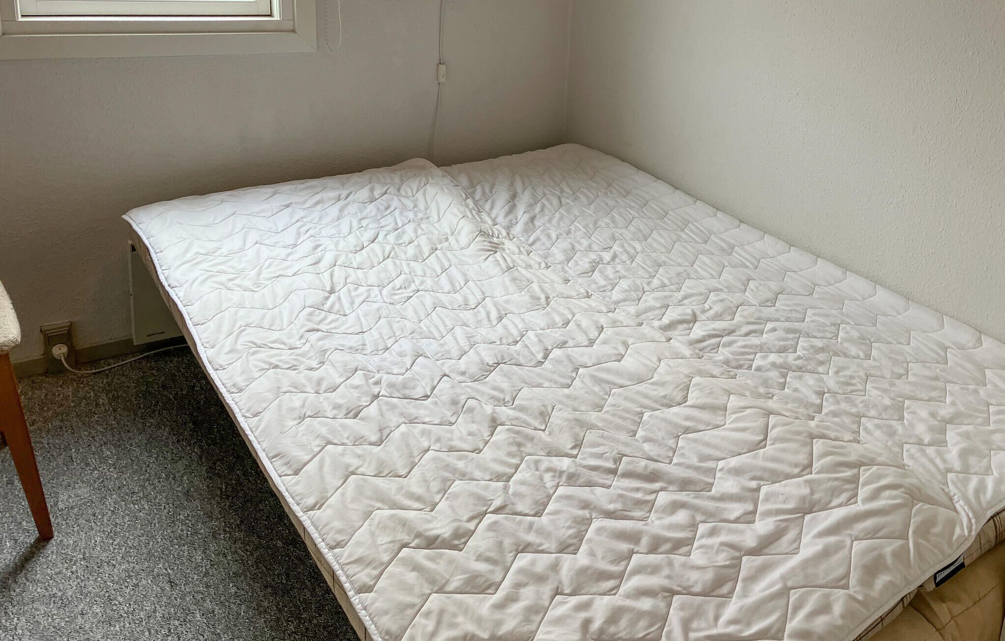 Una camera piccola con un letto singolo, un materasso bianco e una parete semplice e senza decorazioni.