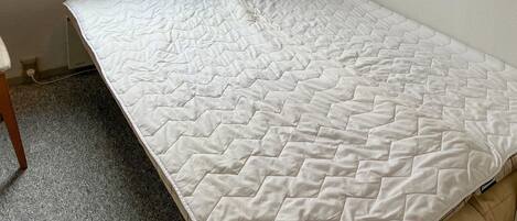 Una habitación pequeña con una cama individual, colchón blanco y una pared sencilla, sin adornos.