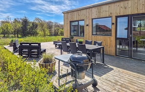 Una terraza de madera con mobiliario exterior, una parrilla y un ventanal grande con vistas a un jardín.
