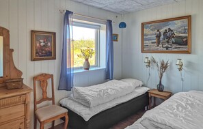 Ein Zimmer mit zwei Betten, einem hölzernen Schminktisch, einem Sessel, einem Fenster mit Vorhängen, Wandleuchten und einem Gemälde.