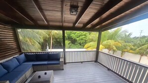 Área de estar coberta ao ar livre com sofá azul e uma mesa pequena, cercada por um corrimão de madeira e vista para palmeiras e uma praia.