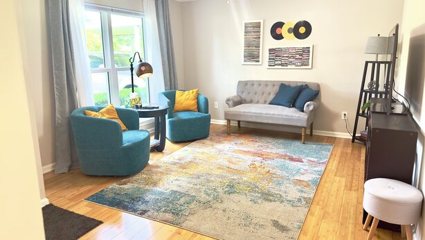 Ein Wohnzimmer mit grauem Sofa, türkisfarbenen Sesseln, einem farbenfrohen Teppich und einem Deckenventilator.