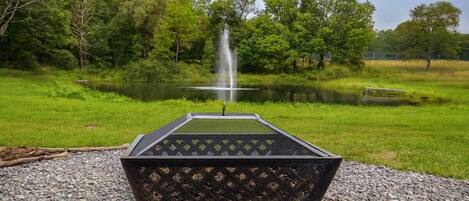 A rectangular outdoor seating area with a metal grill and a view of a pond with a fountain.