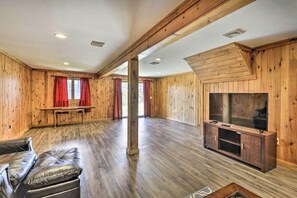 A spacious room with wooden walls and floors, a large window with red curtains, a leather sofa, and a wooden entertainment unit with a television. - Log Cabin and Suite · Stylish Poconos Retreat (Pleasant Mount)