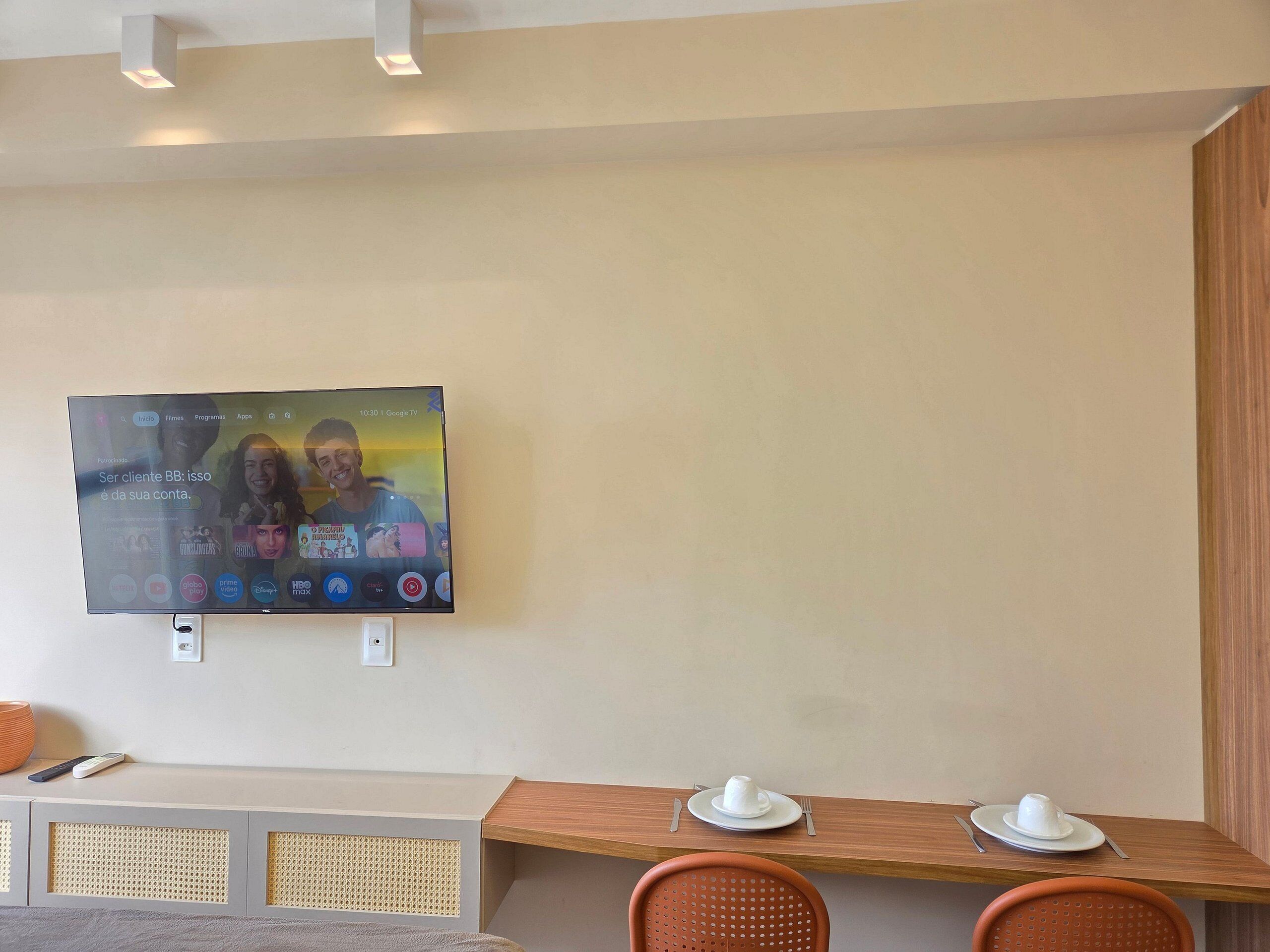 Habitación de hotel con una televisión instalada en la pared mostrando la interfaz de un servicio de streaming. Hay una mesa de madera con un plato blanco y una taza, y dos sillas anaranjadas.