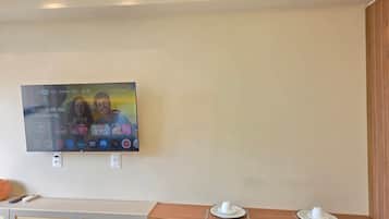 Habitación de hotel con una televisión instalada en la pared mostrando la interfaz de un servicio de streaming. Hay una mesa de madera con un plato blanco y una taza, y dos sillas anaranjadas.