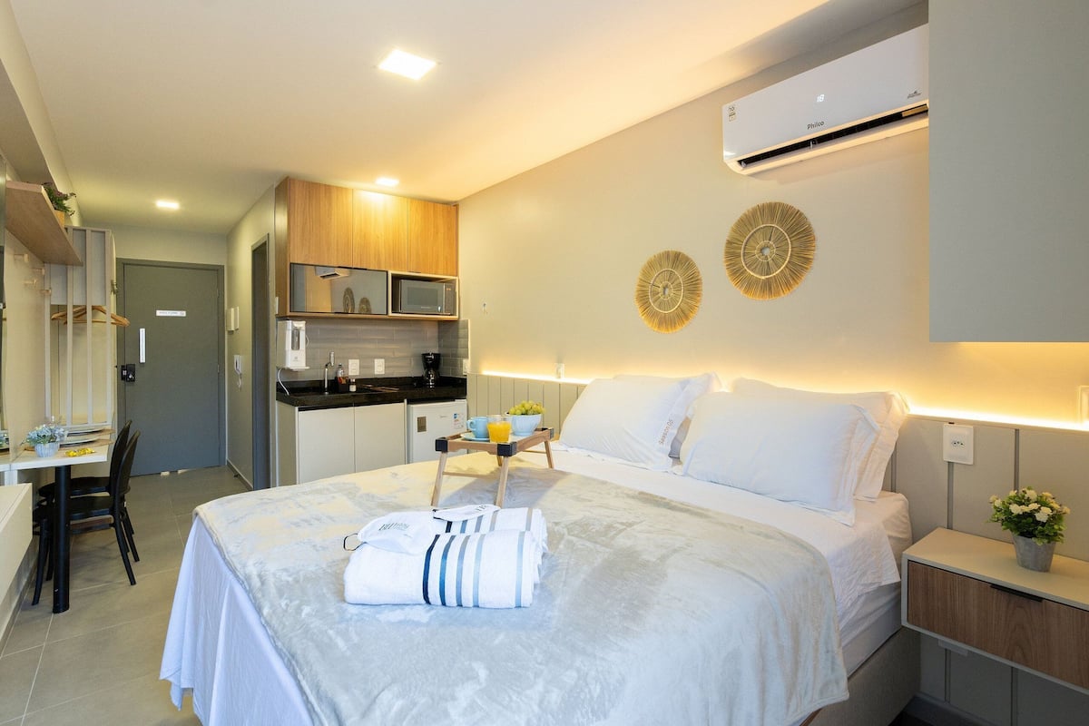 A hotel room with a bed, bedside table, a small desk with a chair, a microwave, and a kitchenette.