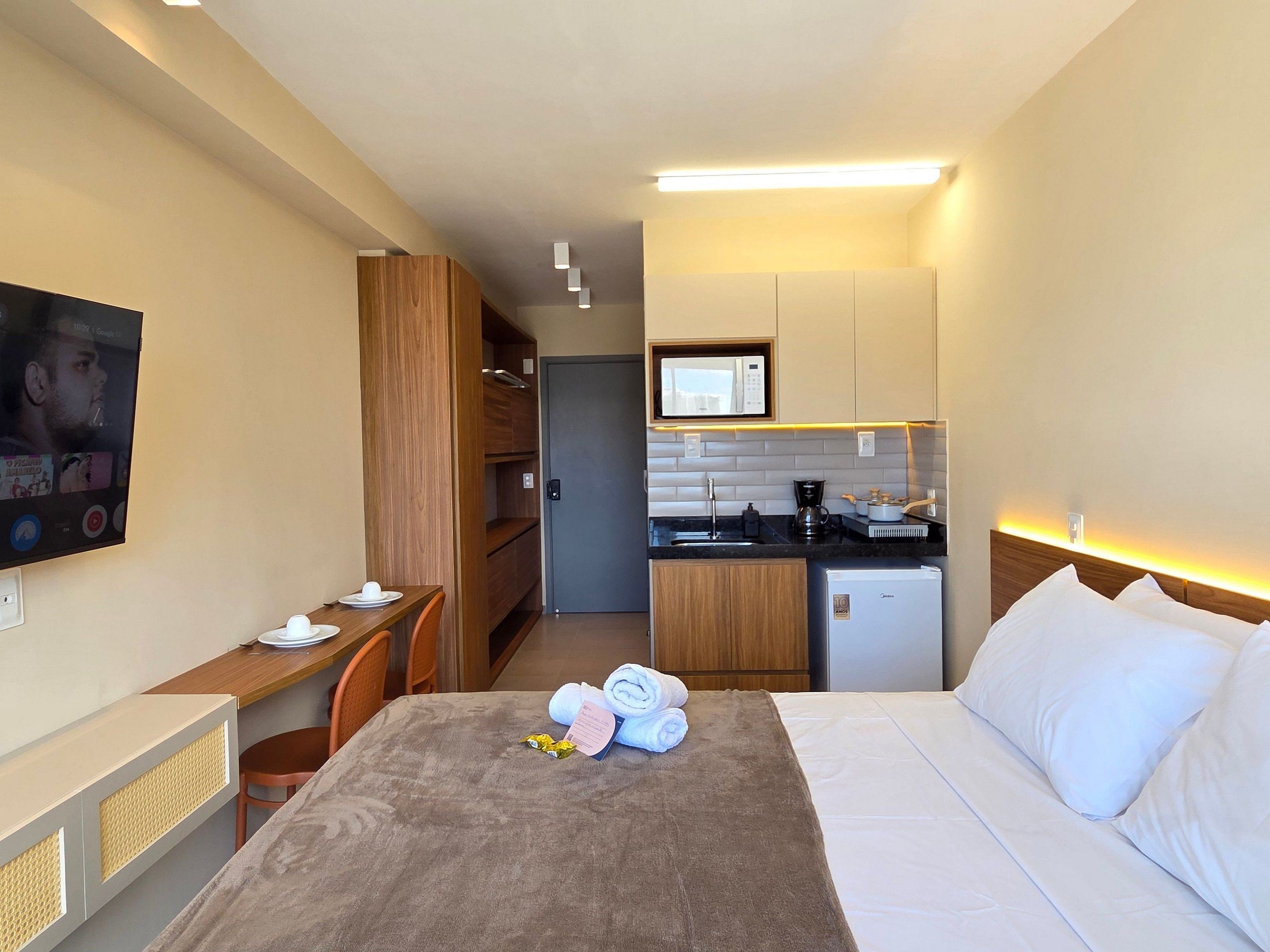 Quarto de hotel com cama, escrivaninha, televisão, cozinha compacta e banheiro.