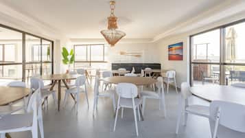 A spacious dining area with white chairs and tables, a hanging light fixture, and large windows.