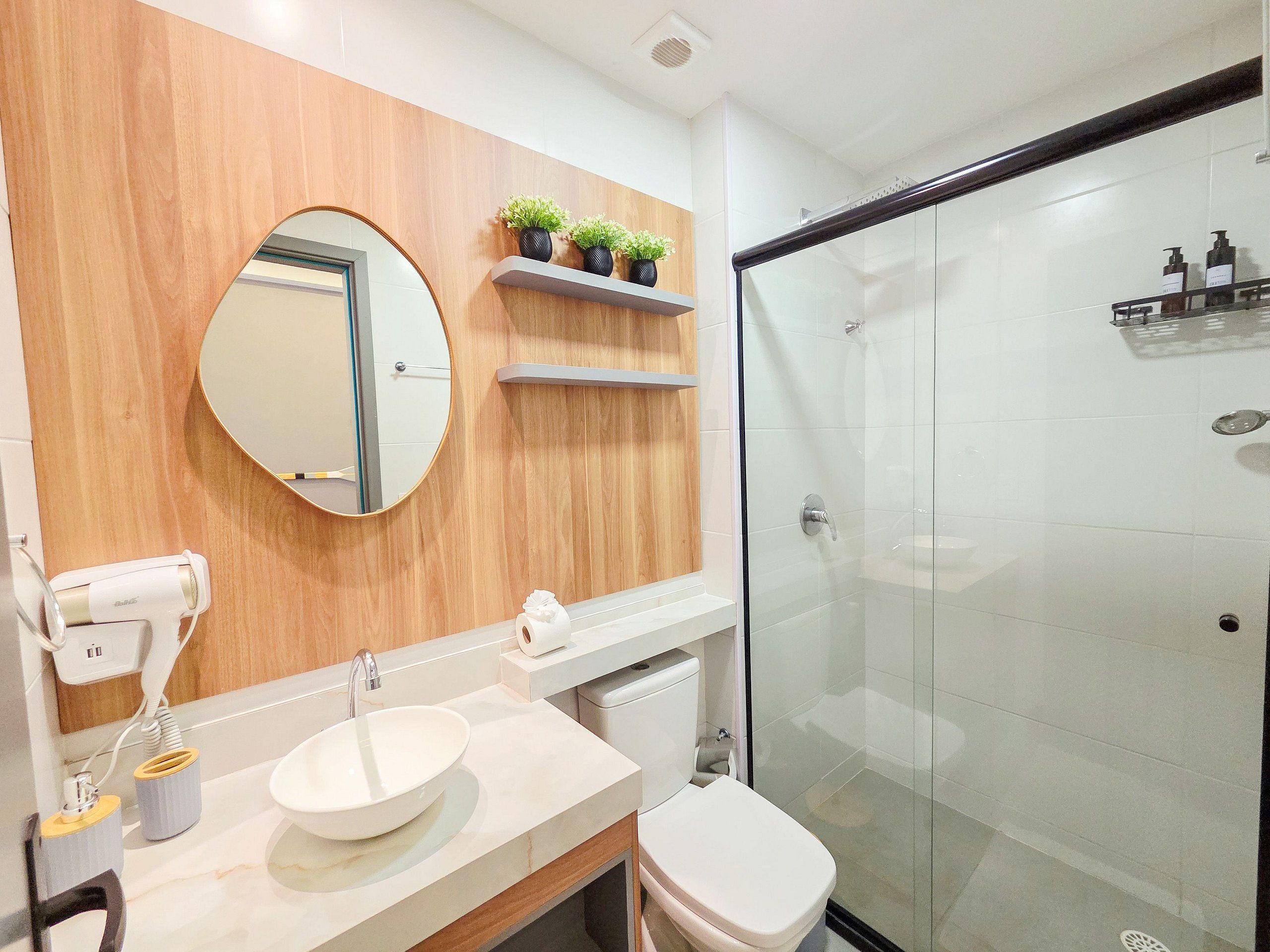 A modern bathroom with a wooden vanity, a round mirror, a toilet, and a glass-enclosed shower.