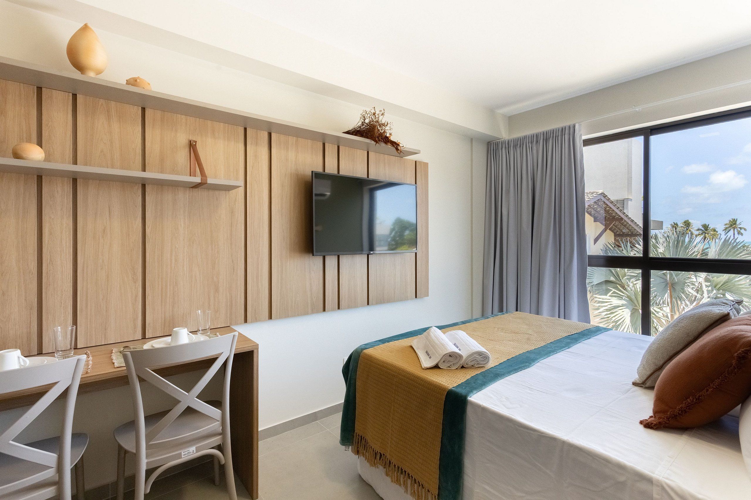 Habitación de hotel con cama, escritorio con sillas, televisor montado en la pared y un gran ventanal con vista a palmeras.