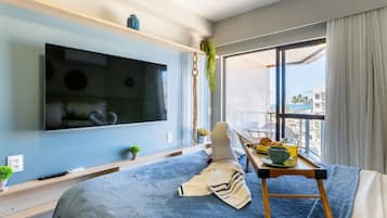 A modern bedroom with a large flat-screen TV mounted on the wall, a bed with a blue blanket, a small table with breakfast items, and a view of the city through large windows.
