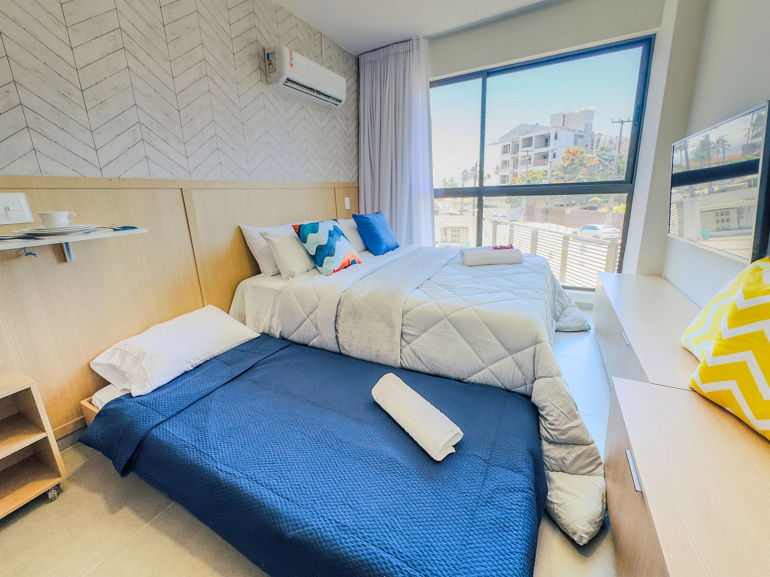 Ett hotellrum med ett stort fönster, en säng med blått sänglinne, en andra säng med vitt sänglinne, en sänggavel i trä och en väggmonterad luftkonditioneringsanläggning.