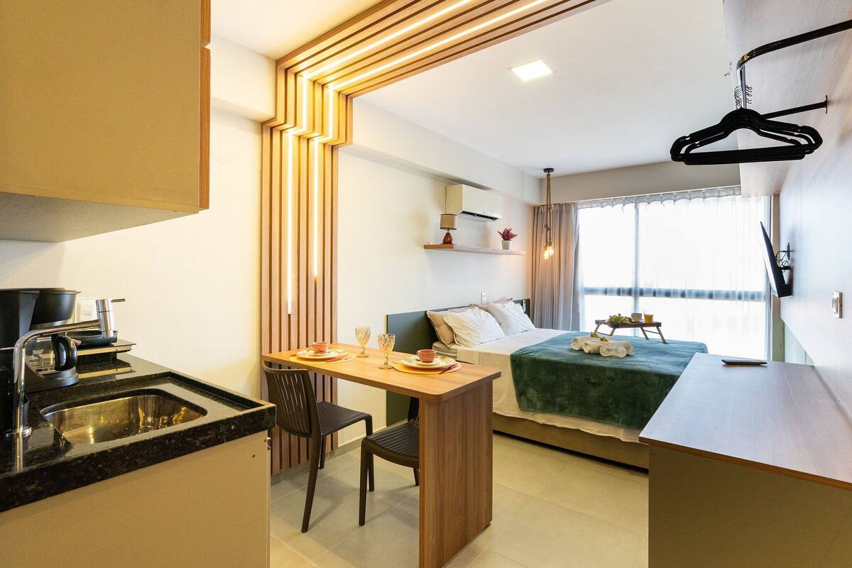 A compact hotel room with a kitchenette, a bed with a green blanket, a bedside table with a lamp, and a window with curtains.