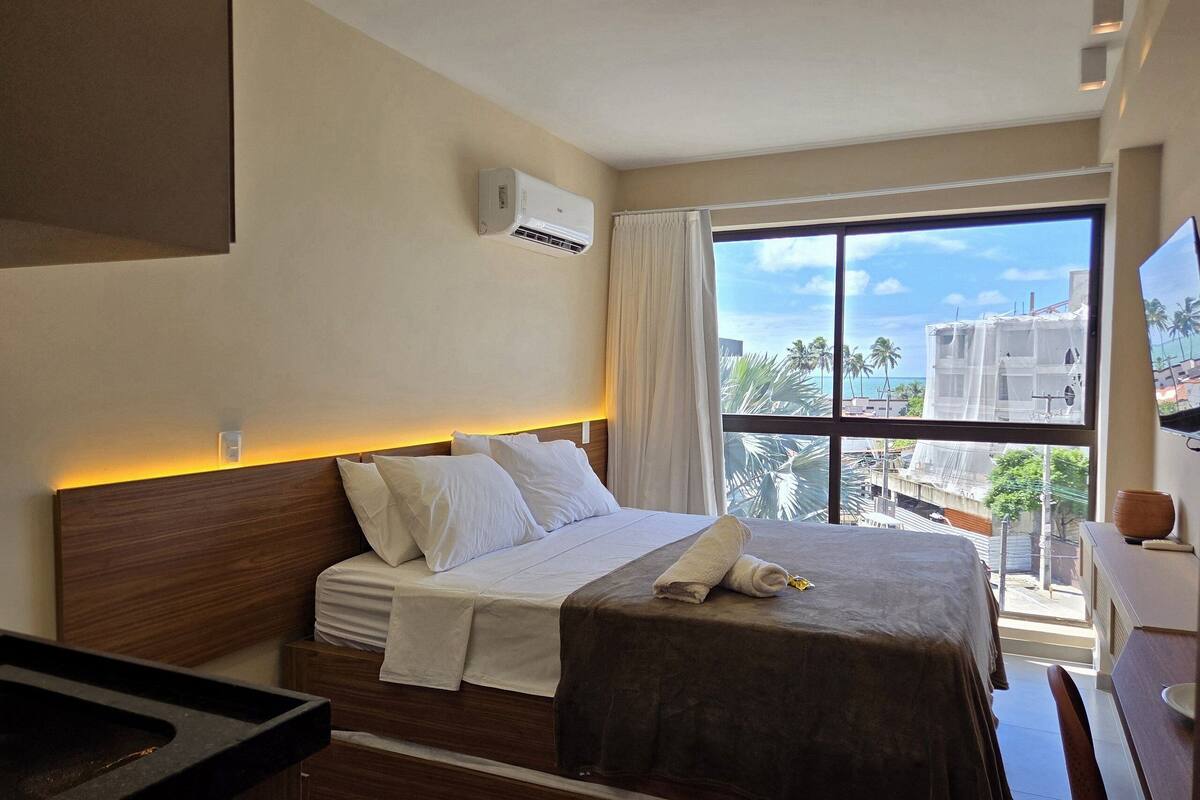 A hotel room with a bed, a window with a view of palm trees and buildings, a wall-mounted air conditioner, and a bedside table.