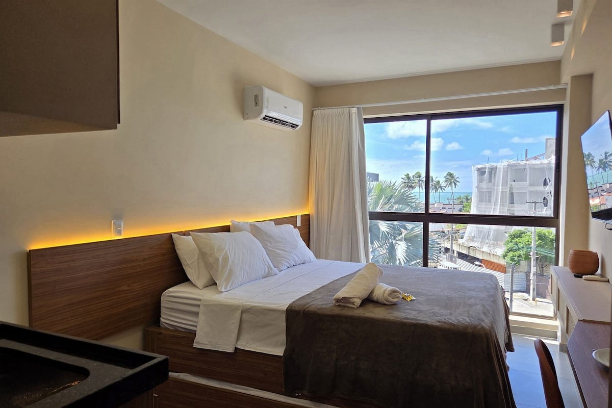 A hotel room with a bed, a window with a view of palm trees and buildings, a wall-mounted air conditioner, and a bedside table.