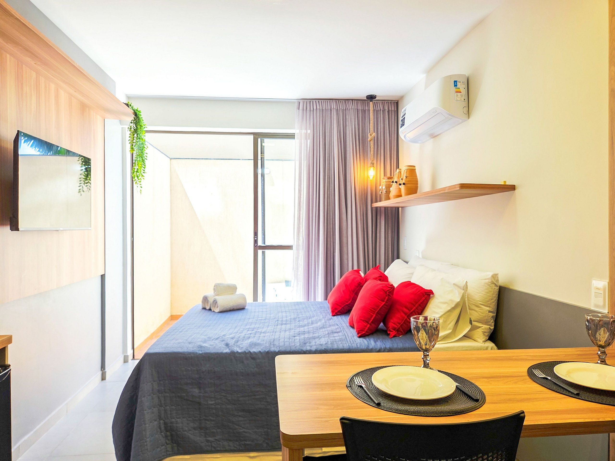 A modern hotel room with a bed, a dining table with a glass, and a wall-mounted air conditioner.