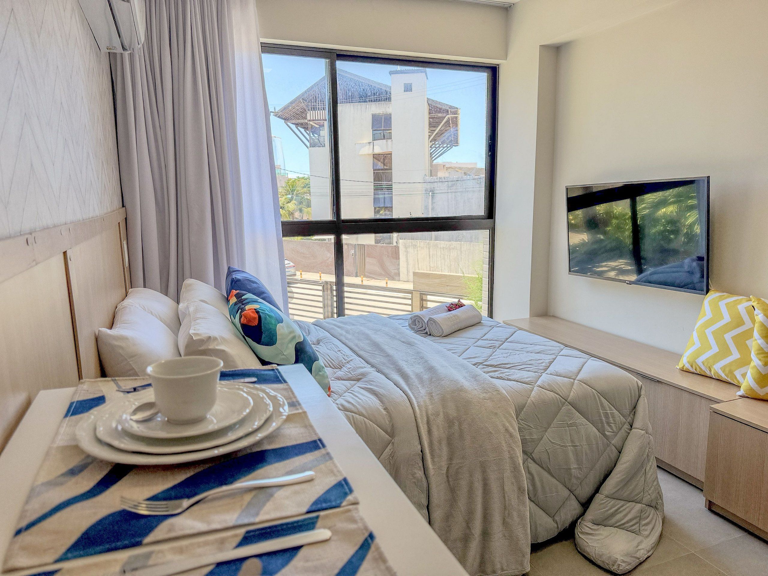 Ein Hotelzimmer mit einem Bett, einem Fenster mit Blick auf Gebäude, einem Fernseher an der Wand und einem Tisch mit Tellern und Besteck.