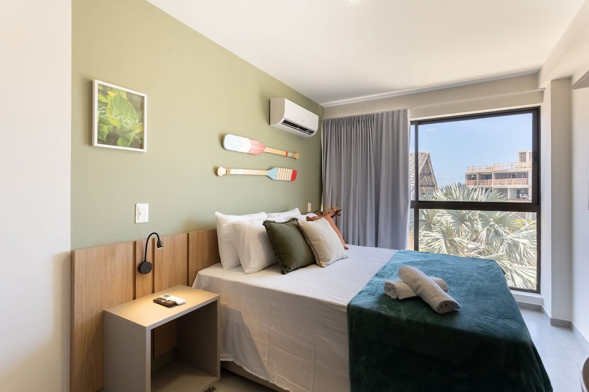A hotel room with a bed, a desk, a window with a view, and a wall-mounted air conditioner.