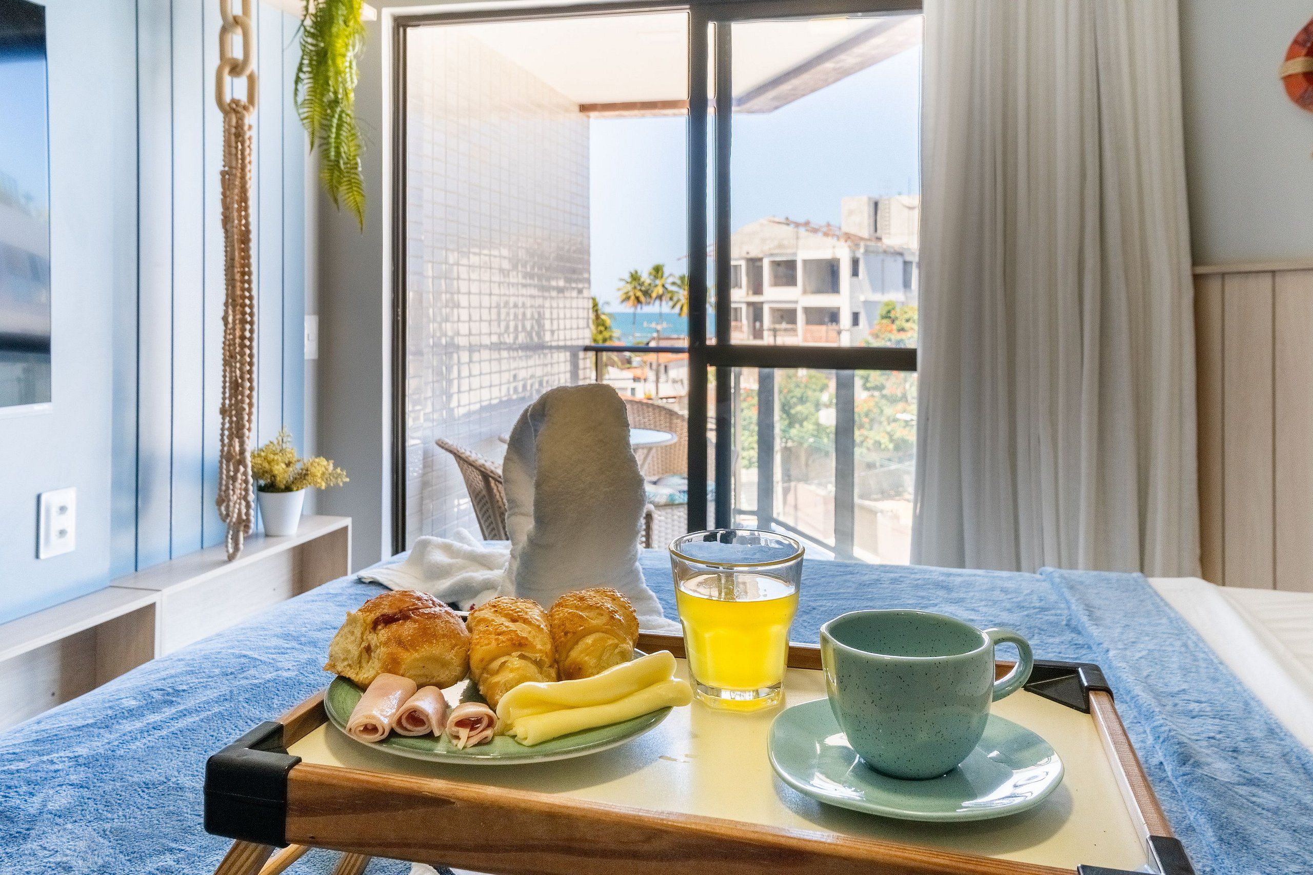 Bandeja de café da manhã com pães, presunto e um copo de suco sobre uma mesa perto de uma janela com vista para edifícios.