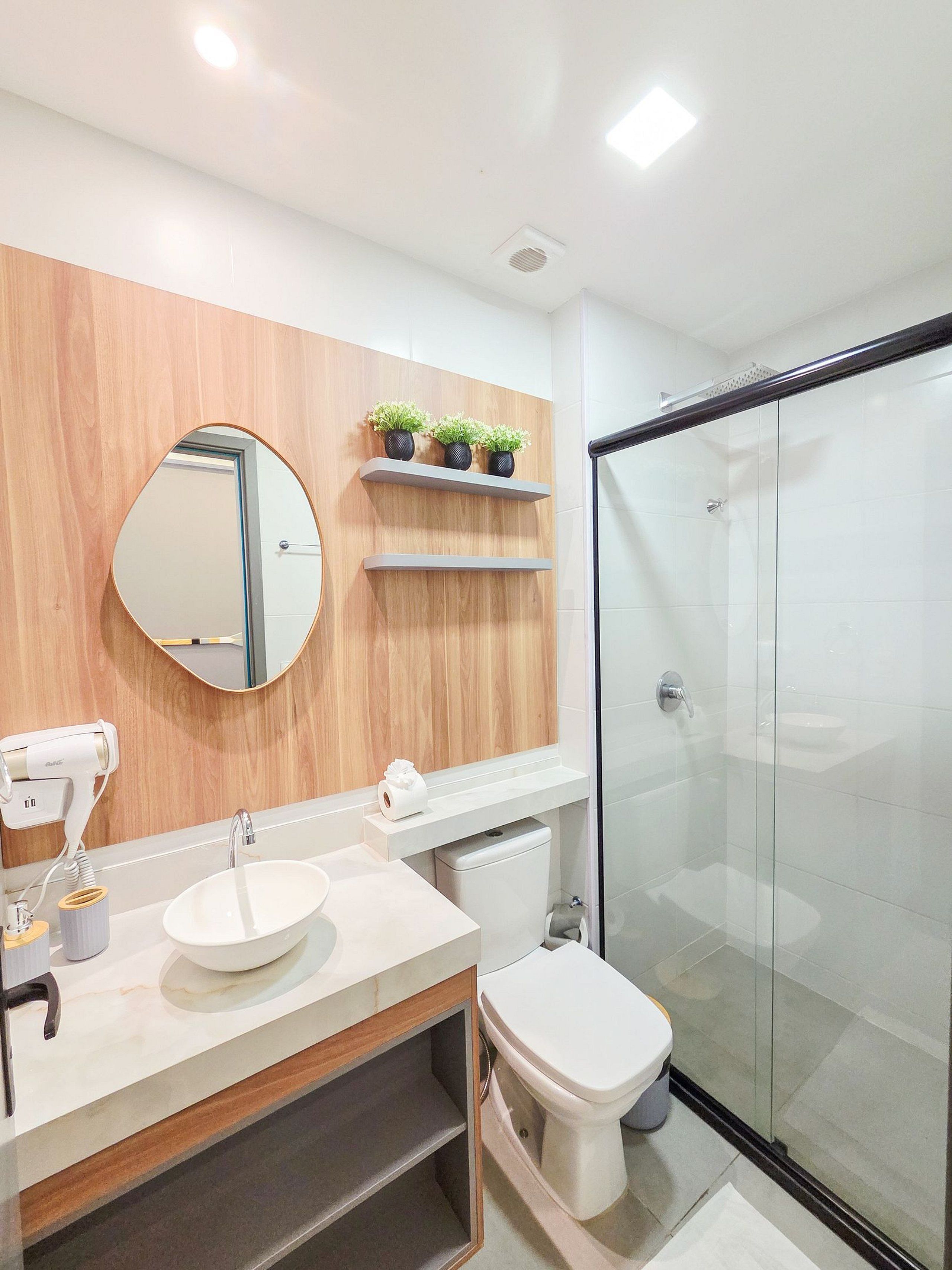 A modern bathroom with a wooden vanity, a round mirror, a toilet, and a glass-enclosed shower.