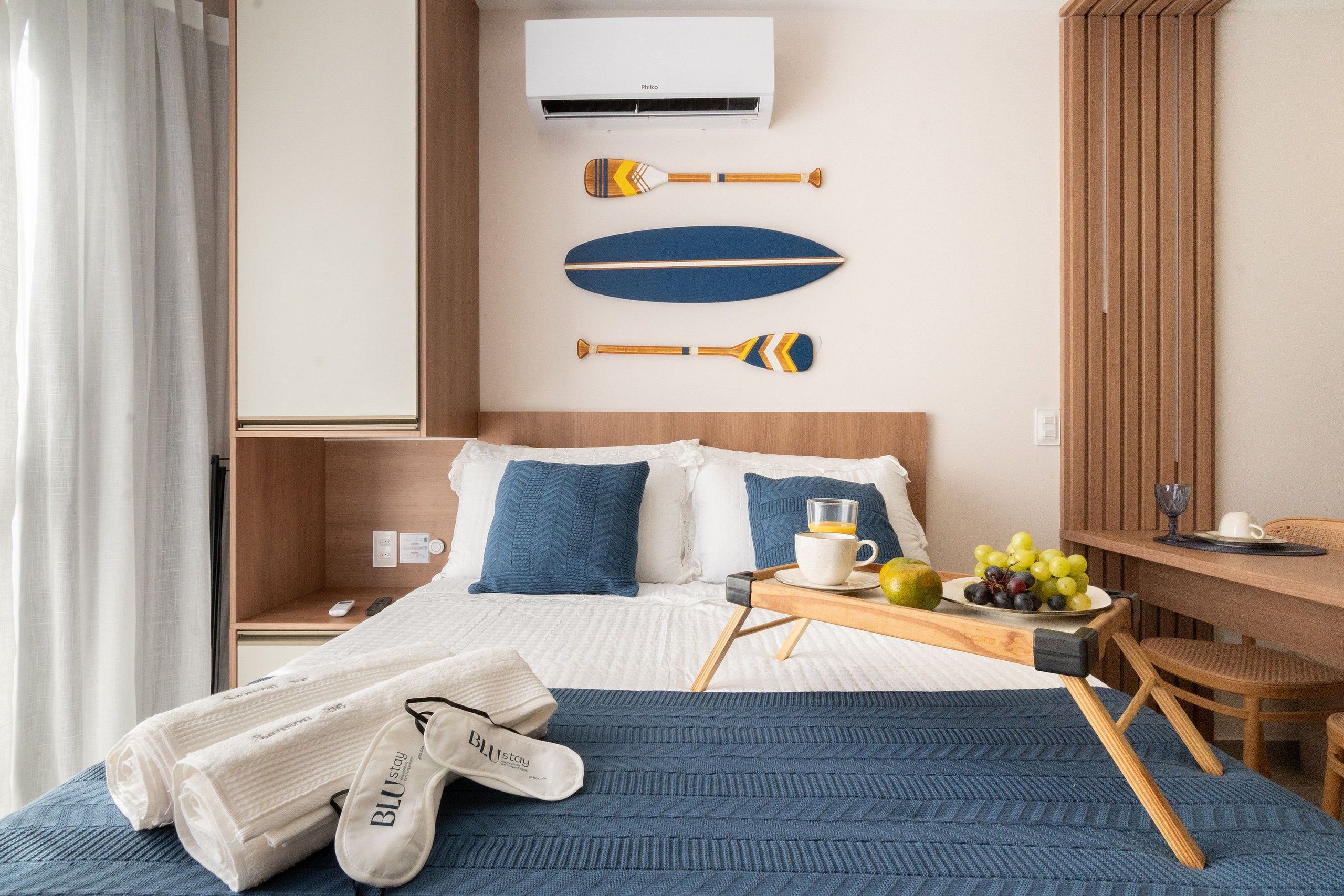 Quarto de hotel com cama, mesa de cabeceira com uma chávena e fruta, uma cadeira e ar condicionado fixado na parede.