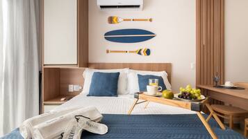 Quarto de hotel com cama, mesa de cabeceira com uma chávena e fruta, uma cadeira e ar condicionado fixado na parede.
