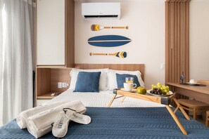 A hotel room with a bed, a bedside table with a cup and fruit, a chair, and a wall-mounted air conditioner. - SEASON Smart Flats by BLU stay (Ipojuca)