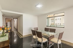 A dining area with a glass table and wicker chairs, a vase of flowers, and a window with a view of buildings.