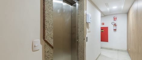 Un ascensor con pantalla digital, un extintor y una puerta roja de emergencia en un pasillo.