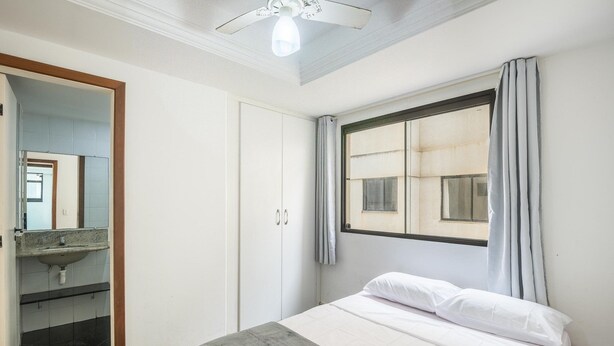 A bedroom with a bed, a window with curtains, a ceiling fan, and a bathroom with a sink and mirror.