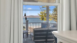 A view from a window with a wooden ceiling and a ceiling fan, overlooking a deck with outdoor furniture and a body of water in the distance. - Modern-coastal retreat with lake views, covered deck, and walkable downtown Beulah location. (Beulah)