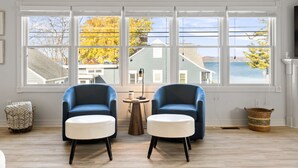 A room with two blue armchairs and white ottomans, a small round table, and a view of a lake and houses through large windows. - Modern-coastal retreat with lake views, covered deck, and walkable downtown Beulah location. (Beulah)