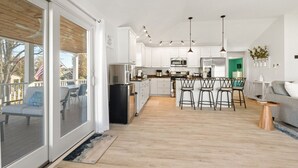 A modern kitchen with white cabinets, a central island with bar stools, a dining area with a green accent wall, and a sliding glass door leading to a deck. - Modern-coastal retreat with lake views, covered deck, and walkable downtown Beulah location. (Beulah)