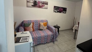 A compact living space with a sofa, a small table, and a refrigerator. There are two wall-mounted pictures, one of a flower and another of a rock arrangement. - Apartment° Fiumicino Rosy 3 (Isola Sacra)