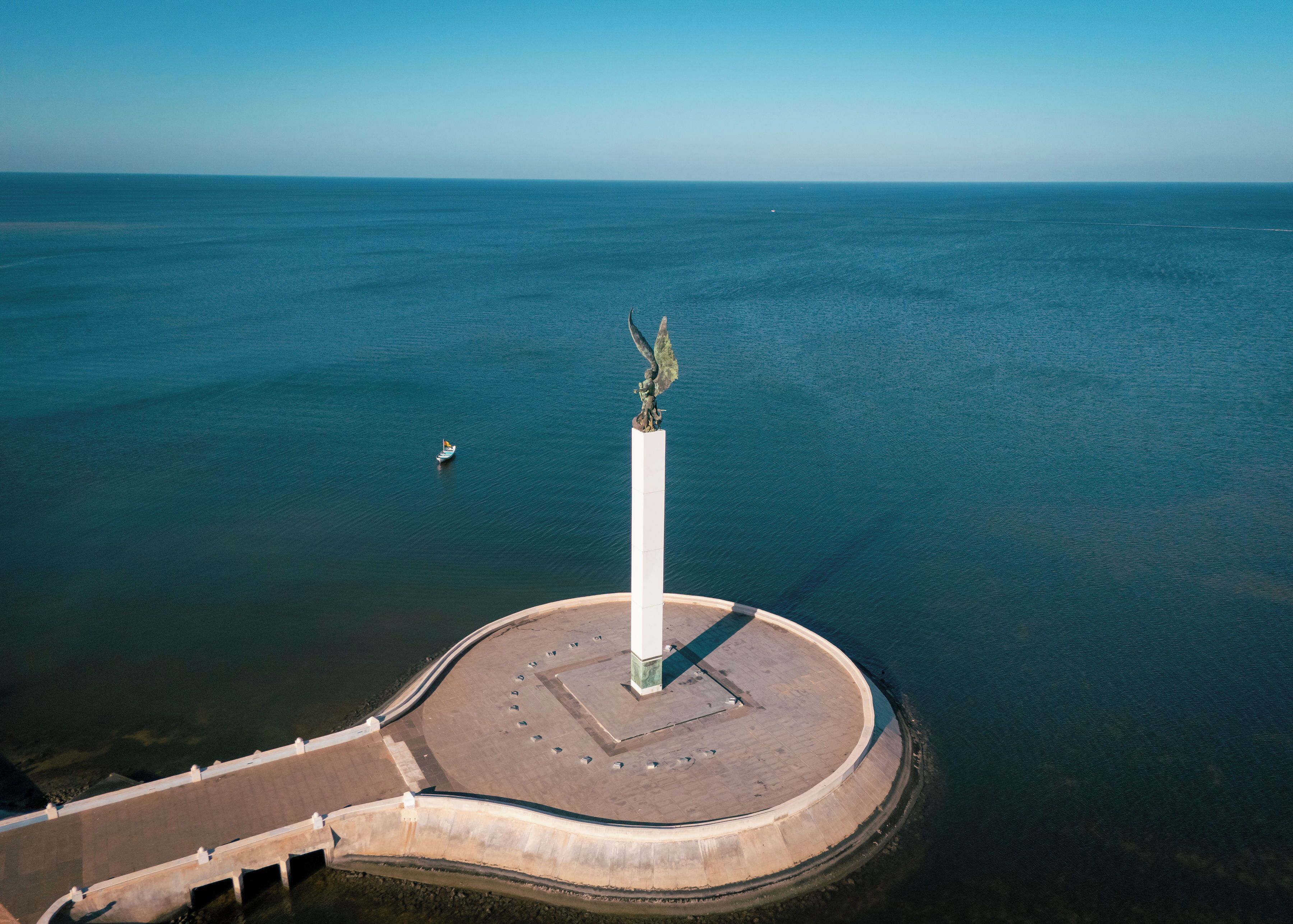 Vista aérea de un paseo marítimo con un monumento central, una plaza circular y un mar tranquilo.