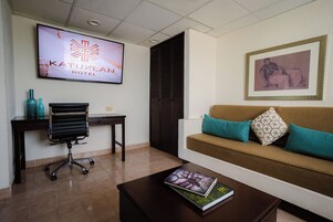 Habitación de hotel con una televisión mostrando el nombre del hotel, un sofá con cojines color turquesa, una mesa de centro con una revista, un escritorio con una silla y una obra de arte enmarcada en la pared.