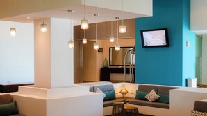 Un moderno vestíbulo de hotel con una pared de acento color verde azulado, un televisor de pantalla plana grande y una zona de estar con sofá y lámpara.