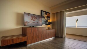 Zona de recepción de un hotel con un mostrador de madera, un televisor mostrando el nombre del hotel y un cuadro abstracto enmarcado.