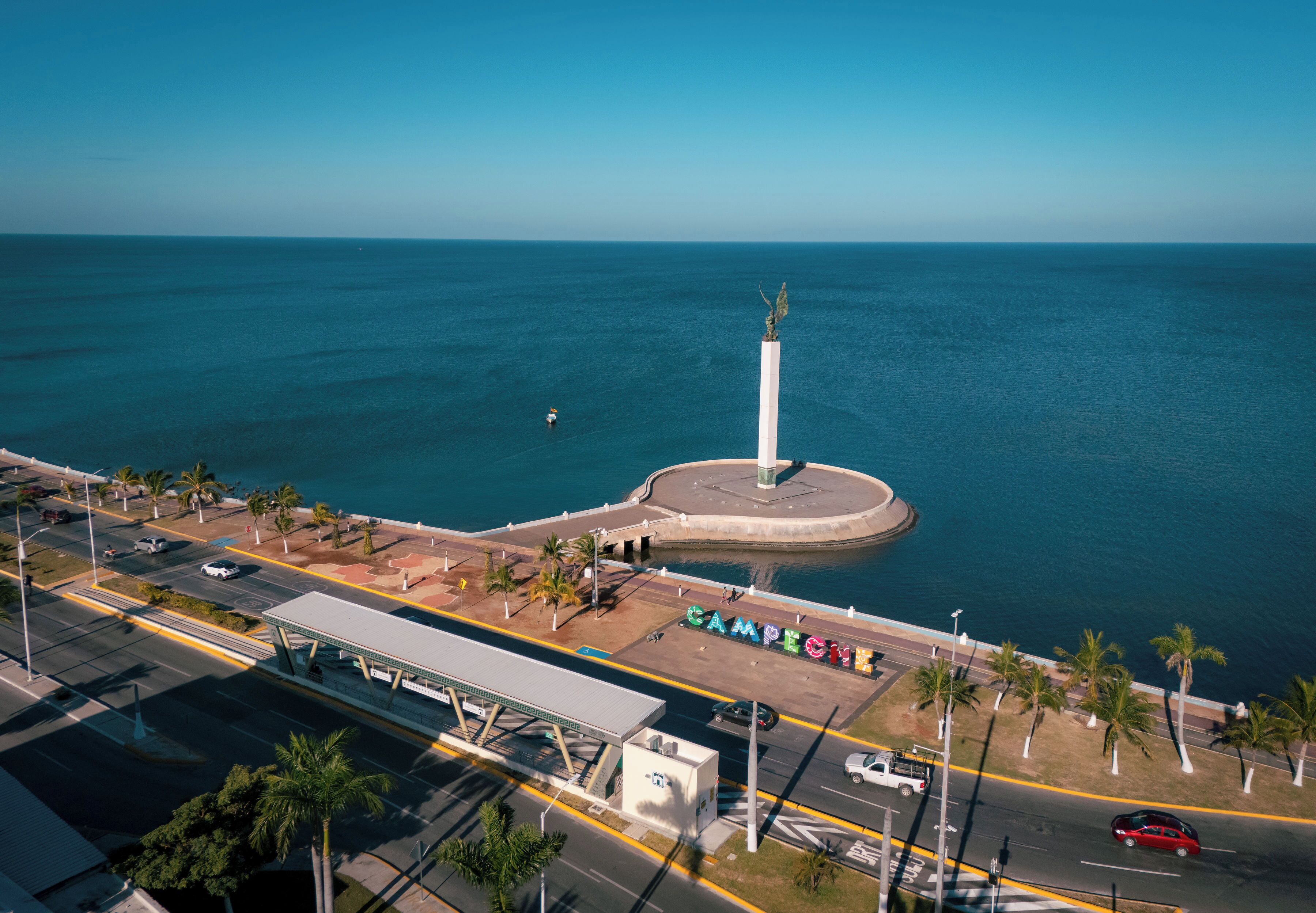 An aerial view of a coastal area with a prominent white monument, a road with vehicles, and a parking lot with the word 'CAMPECIO' visible.