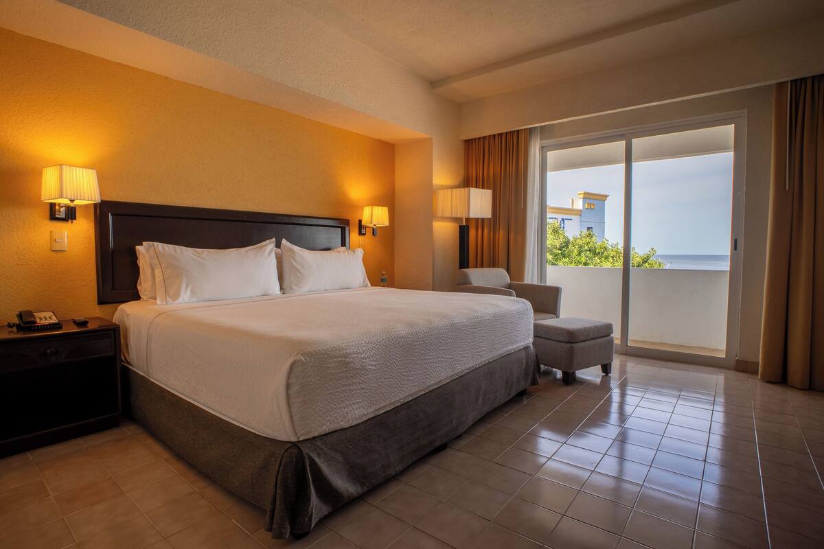 A hotel room with a large bed, two bedside lamps, a nightstand, a small chair, and a view of the ocean through a sliding glass door.