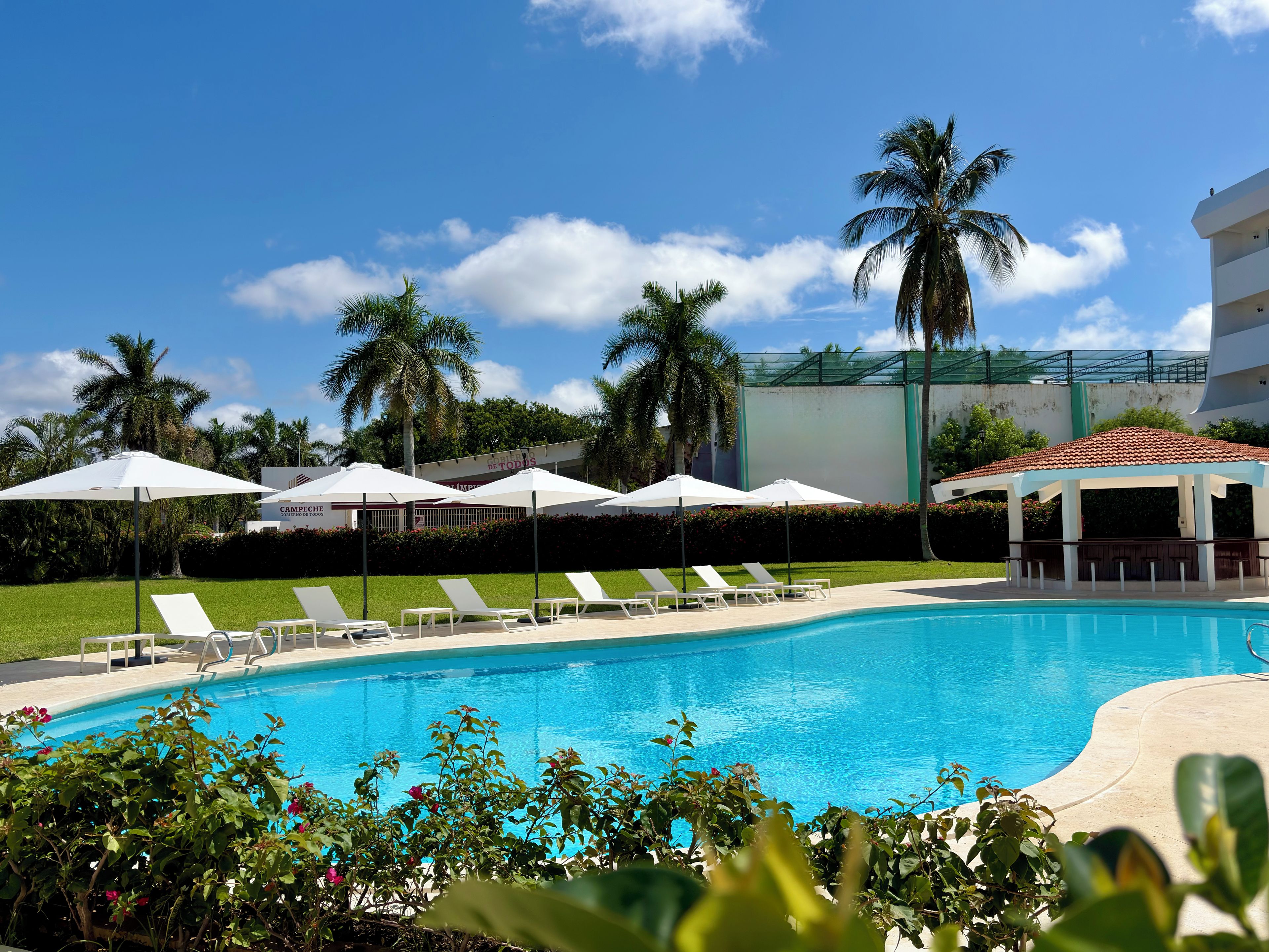 Une piscine entourée de chaises longues et de parasols, bordée de palmiers et d’un bâtiment sur lequel on peut voir une enseigne.