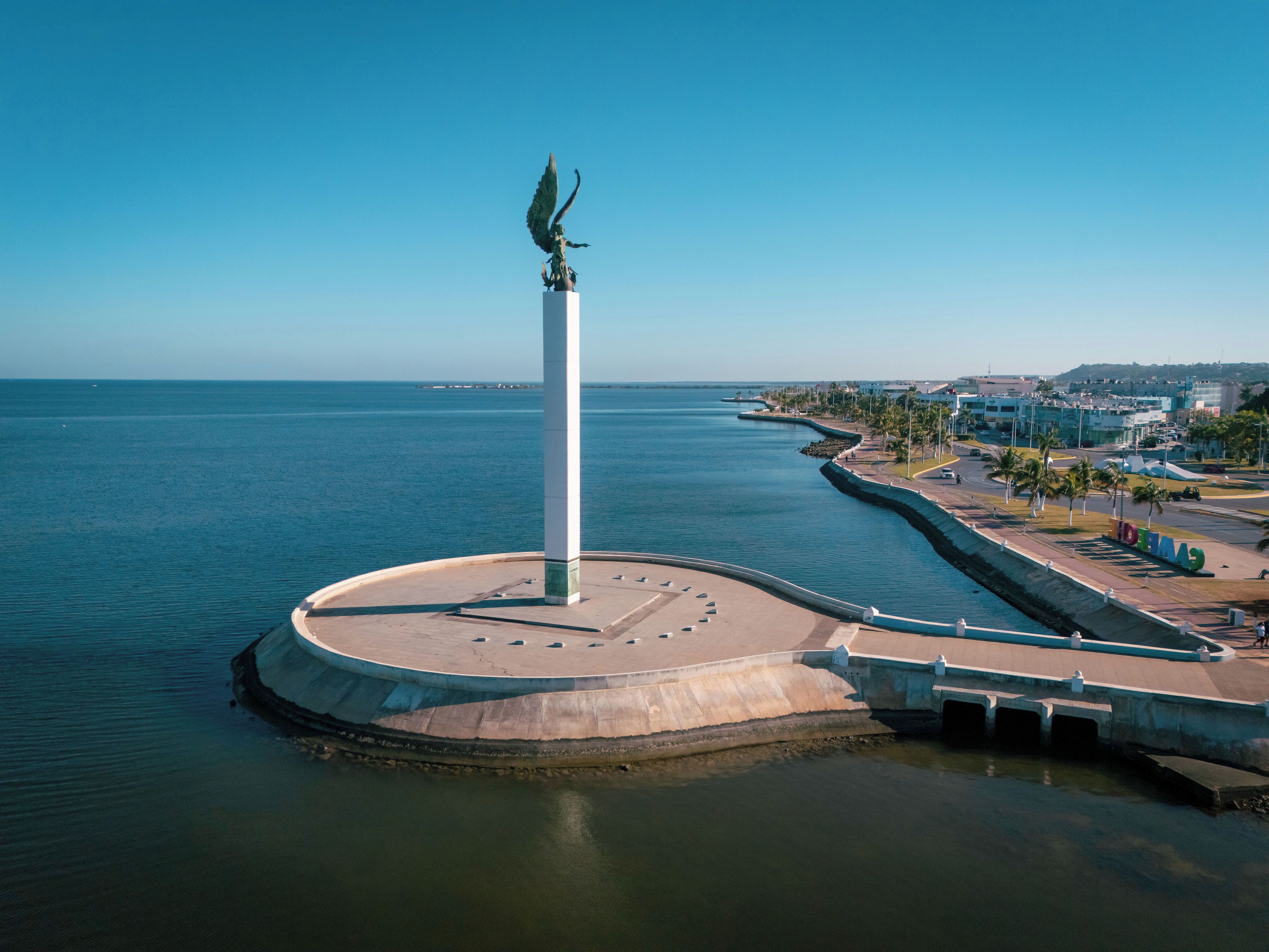 A circular plaza with a central monument, a waterfront promenade, and a distant cityscape.