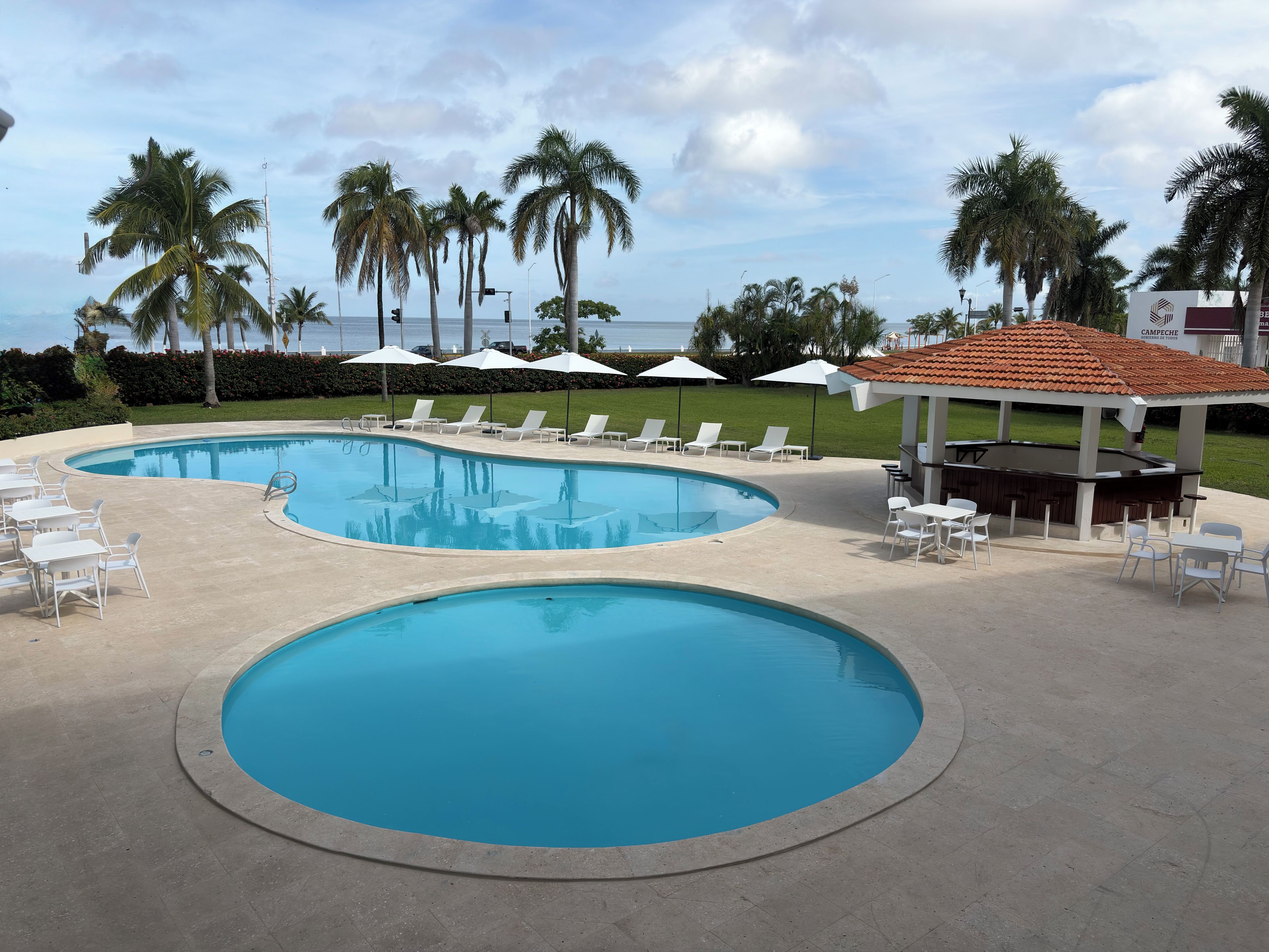 A swimming pool with lounge chairs and umbrellas, surrounded by palm trees and a view of the ocean.